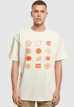 Cremefarvet t-shirt med oversized pasform. Har orange frugtmotiver, herunder citroner og appelsiner, med ordet "SOMMER" i fed skrift nedenfor.
