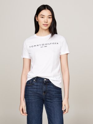 Jeune femme aux longs cheveux noirs portant un t-shirt blanc Tommy Hilfiger et un jean bleu foncé, debout devant un fond uni.