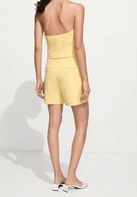 Jumpsuit giallo senza spalline con busto aderente e pantaloni corti sartoriali; include tasche posteriori. Abbinato a sandali bianchi con tacco a blocco.