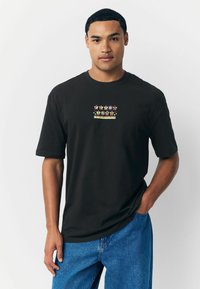 Schwarzes Baumwoll-T-Shirt mit kurzen Ärmeln, das einen bunten Grafikdruck auf der Brust aufweist. Getragen mit blauen Jeans.