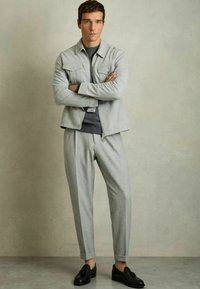 Jachetă de culoare gri deschis, din suede, cu două buzunare la piept, purtată peste un pulover gri închis, asortată cu pantaloni plisati gri deschis și pantofi negri.