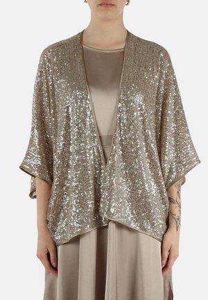Donna che indossa uno shrug aperto davanti con paillettes dorate sopra un vestito in raso taupe, mostrando un tatuaggio floreale sull'avambraccio.