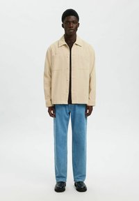 Ung mand står iført en beige knap-opsat jakke, mørk skjorte, lyseblå jeans og sorte sko foran en ensfarvet lys baggrund.