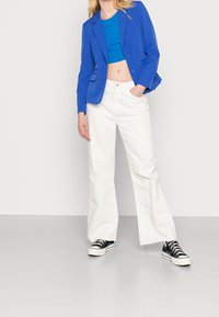 Blazer bleu sur un crop top bleu ajusté, associé à un pantalon large blanc et des baskets à plateforme noires. Tissu lisse, coupe sur mesure.
