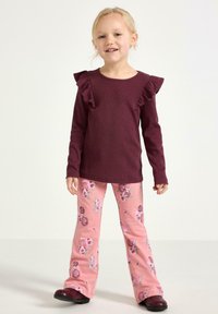 Nuori tyttö seisoo ja hymyilee, pukeutuneena tummanpunainen röyhelöidyllä pitkähihainen paita ja vaaleanpunaiset, kukkakuviolliset housut sekä tummat kengät.