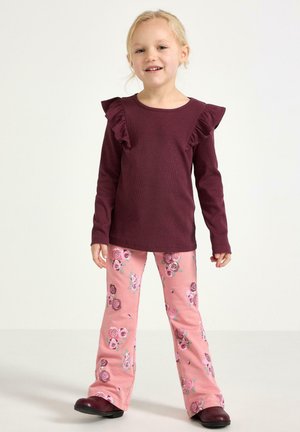 Ung flicka som står och ler, klädd i en vinröd topp med volanger och långa ärmar samt rosa blommönstrade byxor med mörka skor.