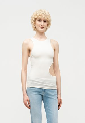 Młoda osoba o kręconych blond włosach, ubrana w biały, bezrękawnik z wycięciem po boku i jasnoniebieskie dżinsy, stojąca na jednolitym tle.