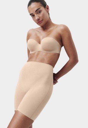 Kvinde iført en beige stropløs bh og beige shaping shorts med høj talje mod en ensartet lys baggrund, poserer med hænderne bag ryggen.