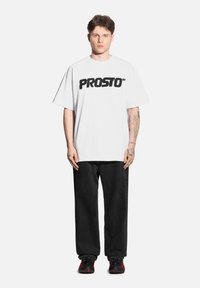 Biała, oversize'owa koszulka z napisem "PROSTO" w odważnym czarnym tekście, w połączeniu z czarnymi, luźnymi spodniami i czarno-czerwonymi sneakerami.
