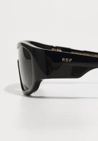 Czarne okulary przeciwsłoneczne z grubymi, błyszczącymi oprawkami, ciemnymi szkłami i złotym logo "RSF" z boku. Zaokrąglone krawędzie i elegancki design.