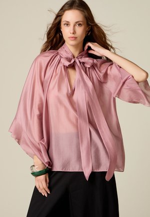 Junge Frau trägt eine transparente rosa Bluse mit einer Schleifenband-Kragen und schwarze Hose, berührt mit einer Hand ihr Haar.