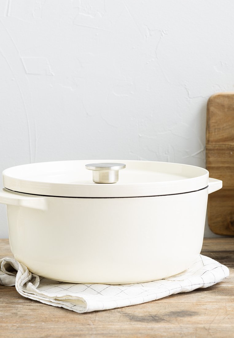 Weißer keramischer Dutch Oven mit Deckel und Metallknopf steht auf einem gefalteten karierten Tuch auf einer Holzarbeitsplatte.