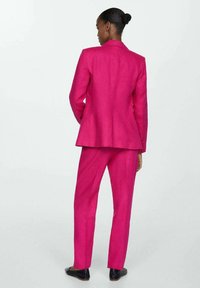 Costum fucsia, ajustat, cu o jachetă structurată și pantaloni slim-fit. Materialul pare neted, cu un ușor luciu. Pantofii negri completează ținuta.