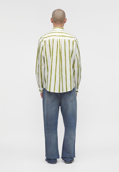 Άτομο με ξυρισμένο κεφάλι που φοράει λευκό πουκάμισο με κατακόρυφες πράσινες ρίγες και φαρδιά μπλε τζιν, βλέπεται από πίσω.
