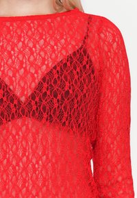 Haut rouge en dentelle à manches longues avec motif floral porté sur un soutien-gorge noir, montrant une texture détaillée et une transparence sur le haut du torse.