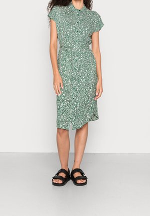 Kvinde iført en grøn og hvid knælange kjole med blomsterprint, korte ærmer, lukket foran med knapper, og sorte åbne sandaler med stropper.