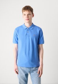 Helles blaues Poloshirt aus weicher Baumwolle, mit Kragen, drei Knöpfen und einem kleinen Logo auf der Brust. Kombiniert mit hellen Jeans.