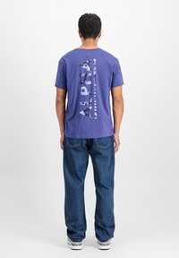 Violeta kokvilnas T-krekls ar vertikāli baltā krāsā drukātu logo uz muguras. Apvienots ar brīviem zilajiem džinsiem. Parasts fons.