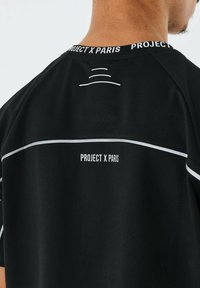 Camiseta negra con líneas blancas y etiqueta "PROJECT X PARIS" en el cuello. Presenta detalles gráficos minimalistas en la parte posterior.