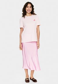Femme portant une chemise à rayures roses et blanches à manches courtes avec des coeurs rouges et une jupe midi en satin rose clair, souriante et debout.