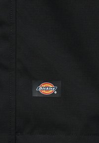 Μαύρο ύφασμα με εμφανή ραφή που διαθέτει ένα μικρό ορθογώνιο ετικέτα με το λογότυπο της μάρκας Dickies σε κόκκινο, κίτρινο, λευκό και ναυτικό μπλε χρώμα.