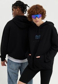 Black hooded sweatshirt with front pocket, featuring blue text on chest. Model wears black shorts and oversized sunglasses. Casual styling.