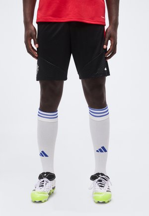 Männlicher Athlet trägt schwarze Shorts, weiße kniehohe Socken mit blauen Streifen und weiße Fußballschuhe mit neongrünen Sohlen, steht vor weißem Hintergrund.