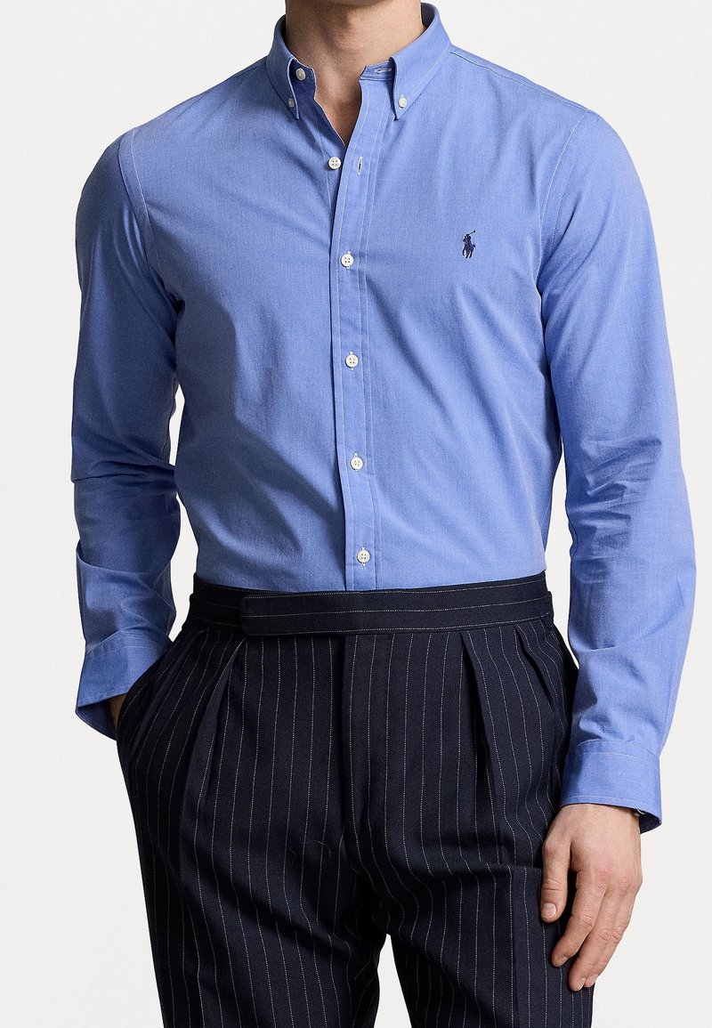 Homme portant une chemise bleu clair boutonnée avec un petit logo sur la poitrine et un pantalon noir à fines rayures, les mains dans les poches.