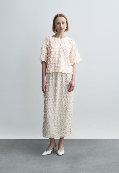 Ροζ ανοιχτόχρωμο τοπ με υφή λουλουδιών και κοντά μανίκια, συνδυασμένο με φούστα μέσου μήκους σε κρεμ απόχρωση με λουλουδάτο μοτίβο και λευκά μυτερά τακούνια.