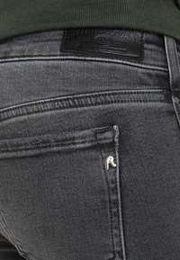 Acercamiento al bolsillo trasero de unos pantalones vaqueros de mezclilla negra con un pequeño detalle metálico en forma de "R" y una etiqueta de cuero en la cintura, combinados con una blusa verde.