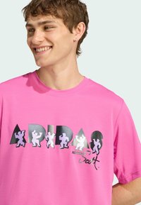Lyserød bomulds t-shirt med iøjnefaldende sort og hvid grafisk tekst "ADIDAS" og små figurer. Kortærmet og rund halsudskæring.
