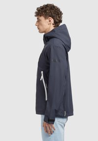 Veste imperméable marine avec capuche, poches zippées et logo KHUOTO. Présente un design élégant et des accents de zip blancs contrastants.