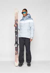 Giacca puffer azzurro chiaro con una striscia bianca, pantaloni da sci neri e scarponi da sci neri. Tiene uno sci decorato con attacchi e occhiali da sole blu.