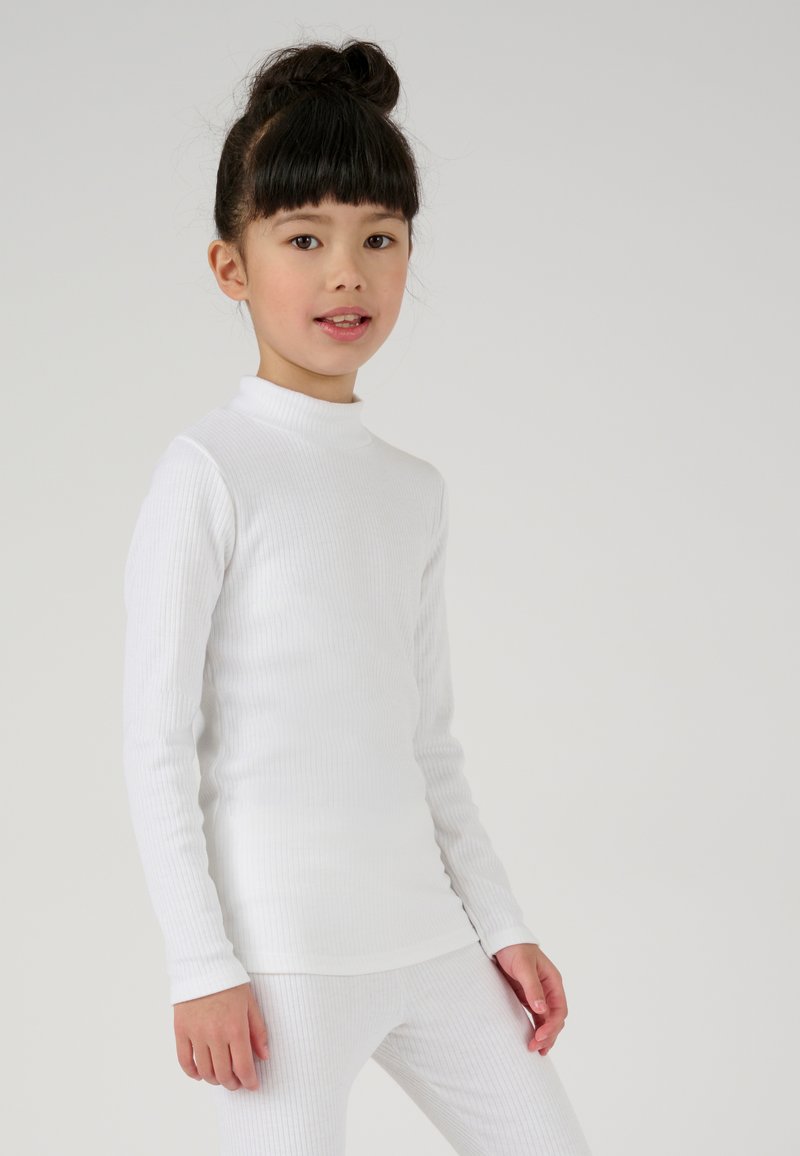 Witte lange mouw top met een hoge kraag en een geribbeld textuur. Gestyled met bijpassende witte geribbelde leggings. De achtergrond is eenvoudig.