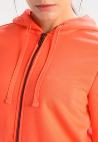 Sweat à capuche corail vif, fabriqué en tissu doux. Il présente une fermeture éclair noire, des cordons de serrage sur la capuche et une texture lisse.