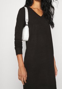 Robe noire tricotée avec un décolleté en V et des manches longues, assortie d'un petit sac à main en croco blanc embossé. Les détails incluent des poignets côtelés.