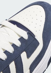 Zapatillas blancas y marino que presentan cuero liso, detalles de ante, puntera perforada y suela de goma texturizada con cordones blancos.