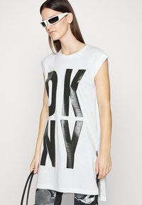 Balta per didelį t-shirt su ryškiu juodu užrašu "OK NY", trumpomis rankovėmis ir šoniniais įpjovimais. Suderinta su juodai baltais raštuotais apatiniais ir saulės akiniais.