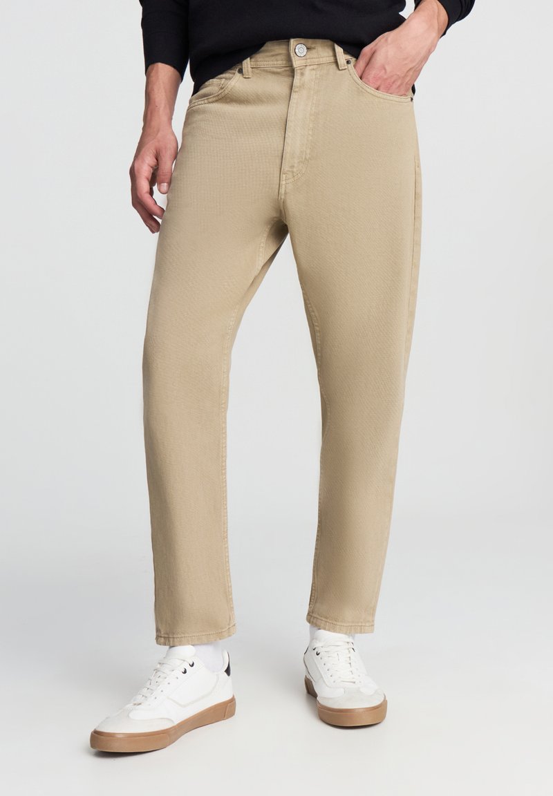 Jeans beige con una texture liscia e design a gamba dritta, dotati di tasche frontali e chiusura con bottone. Abbinati a sneakers bianche.