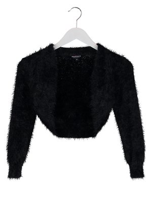 Femme en robe grise, boléro noir en matière duveteuse, collants noirs et talons, tenant une pochette noire devant un fond neutre.