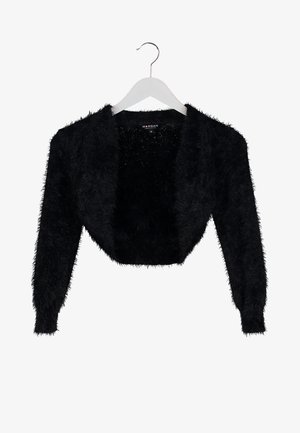 Vrouw in een grijze jurk, zwarte pluizige shrug, zwarte panty's en hakken, met een zwarte clutch tegen een neutrale achtergrond.