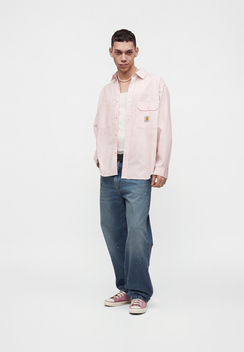 Man wearing light pink button-up shirt over white tank top, loose blue jeans, pink sneakers, and a pearl necklace, standing on white background.