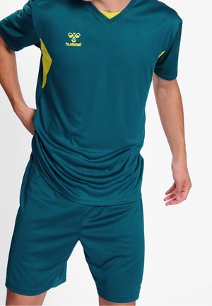 Male wearing teal sportswear with yellow accents, featuring a hummel logo on the chest, posing with one hand on hip and the other relaxed.