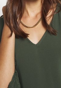 Olivgrünes Oberteil mit V-Ausschnitt und Spitzendetails an den Ärmeln, kombiniert mit einer goldenen Kettenhalskette aus verbundenen Segmenten.