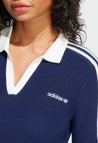 Tamno plava pletena polo majica s bijelom ovratnikom, koja ima dizajn s v-izrezom. Istaknuti bijeli Adidas logo na prsima i tri bijele pruge na rukavima.