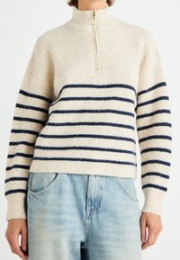 Suéter de punto beige con cuello alto, con franjas horizontales azul marino y un detalle de cremallera. La tela es suave y con textura.