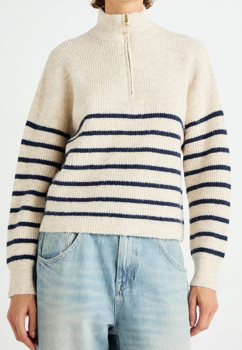 Suéter de punto beige con cuello alto, con franjas horizontales azul marino y un detalle de cremallera. La tela es suave y con textura.
