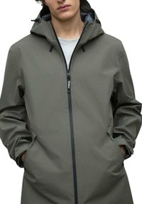 Chaqueta verde resistente al agua con cremallera frontal, capucha ajustable y bolsillos laterales. Presenta una textura suave y un diseño minimalista.