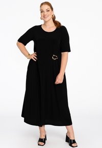 Schwarzes, knielanges Kleid mit kurzen Ärmeln und rundem Ausschnitt, versehen mit einer seitlichen Tasche, die mit einem goldenen Akzent geschmückt ist. Glatte Stoffstruktur.