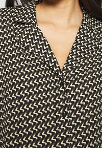 Bluse i sort og creme med zigzag-mønster, krave, knaplukning og glat stof. Nærbillede af halsudskæringen.